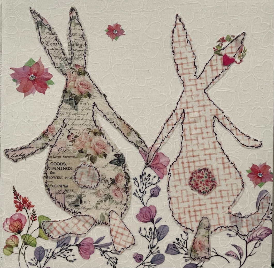Textile bunny art showing two rabbits holding hands with soft floral accents in a gentle, romantic composition