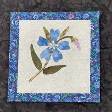 Blue fabric collage flower with crystal center and green leaves on a neutral textile background, mounted on an 8x8 panel