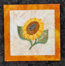 Hand-appliquéd sunflower textile artwork with yellow petals, a beaded center, and layered fabric on a neutral background mounted on an 8x8 panel