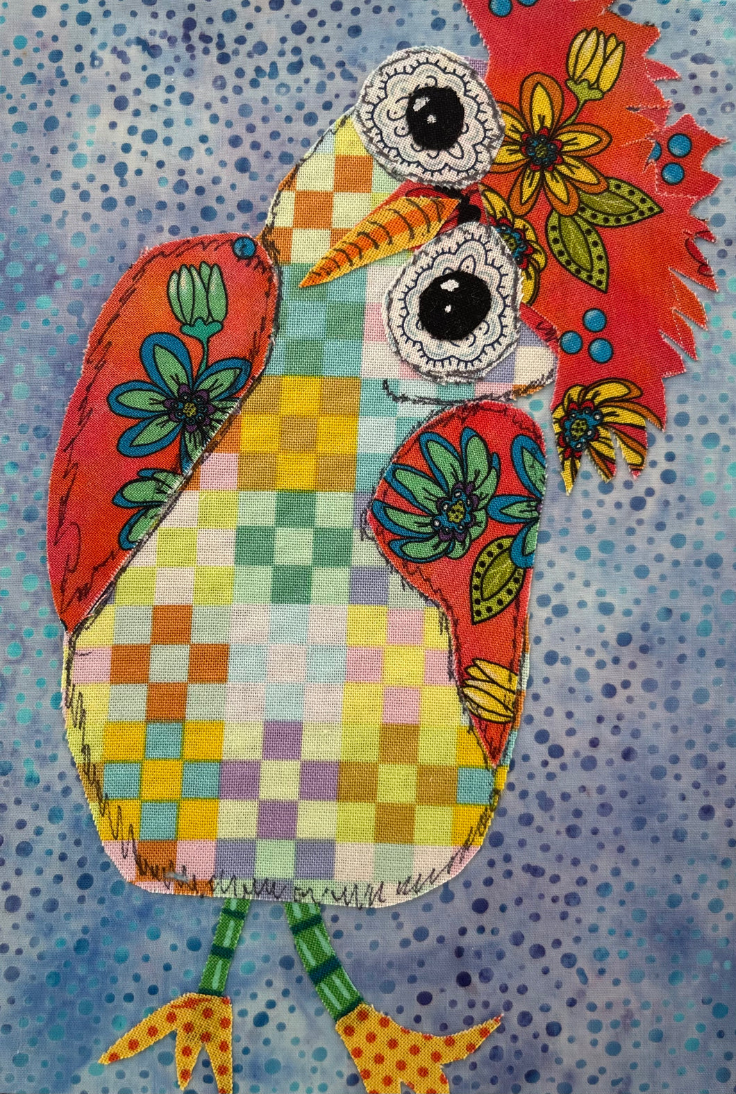 Colorful whimsical fabric collage bird with patchwork body, floral wings, and large expressive eyes on a blue textured background