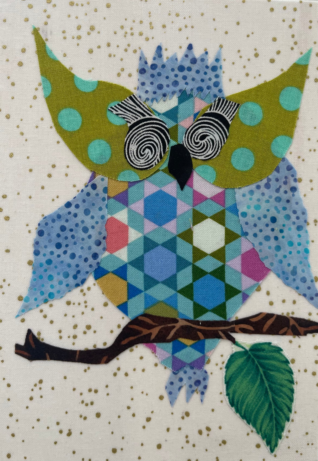 Whimsical fabric collage owl with patterned body, large spiral glasses, and blue dot wings perched on a branch with a leaf on a softly speckled background