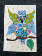 Quirky fabric collage owl with patterned body, large spiral glasses, and blue spotted wings perched on a branch with a leaf on a softly gold speckled background