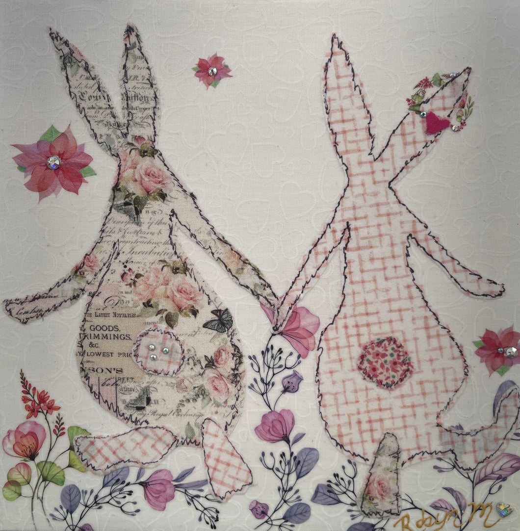 Textile bunny art showing two rabbits holding hands with soft floral accents in a gentle, romantic composition