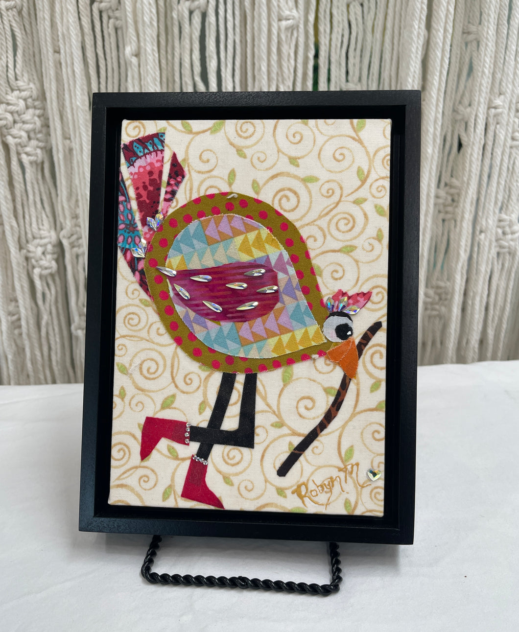 Whimsical fabric collage bird with colorful patterned body holding a walking stick, set against a soft swirling background