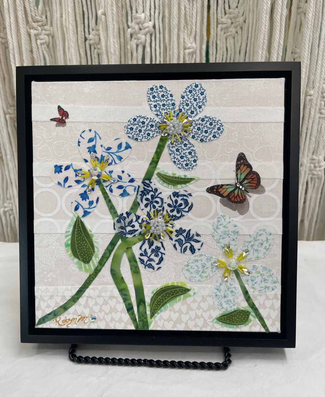 Fabric flower artwork with blue and white patterned blooms, green stems, and butterfly accents on 8x8 panel