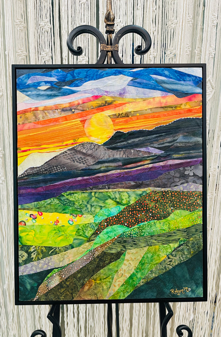 Original all fabric landscape collage artwork 20x16 inches, mountain sunset scene with golden sun descending behind dark mountain silhouettes with Swarovski crystals, fiery orange and red sky, deep cobalt clouds, sweeping landscape layers in violet green and yellow batik fabric with Swarovski crystals, on birch panel, part of the Where Color Takes Root Collection by Robyn Moore Arts
